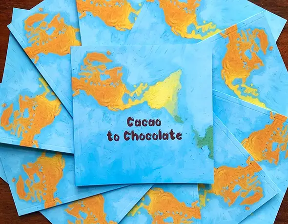 Copies of the 'Cacao to Chocolate' book fanned out on a wooden table, each cover hand-painted as a blue and orange world map.